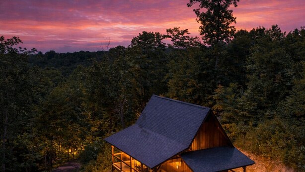 A cabin with a large deck, surrounded by dense forest, at dusk.
