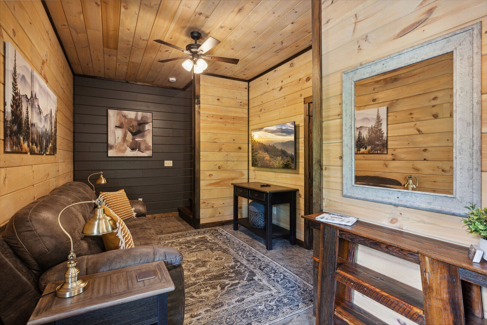 A cozy cabin interior with a leather sofa, a small table with a lamp, a ceiling fan, and framed artwork on the walls.