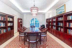 A library with a large wooden table, wicker chairs, and tall bookshelves filled with books.
