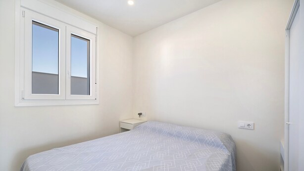 Un dormitorio con cama, ventana, mesita de noche y toma de corriente.