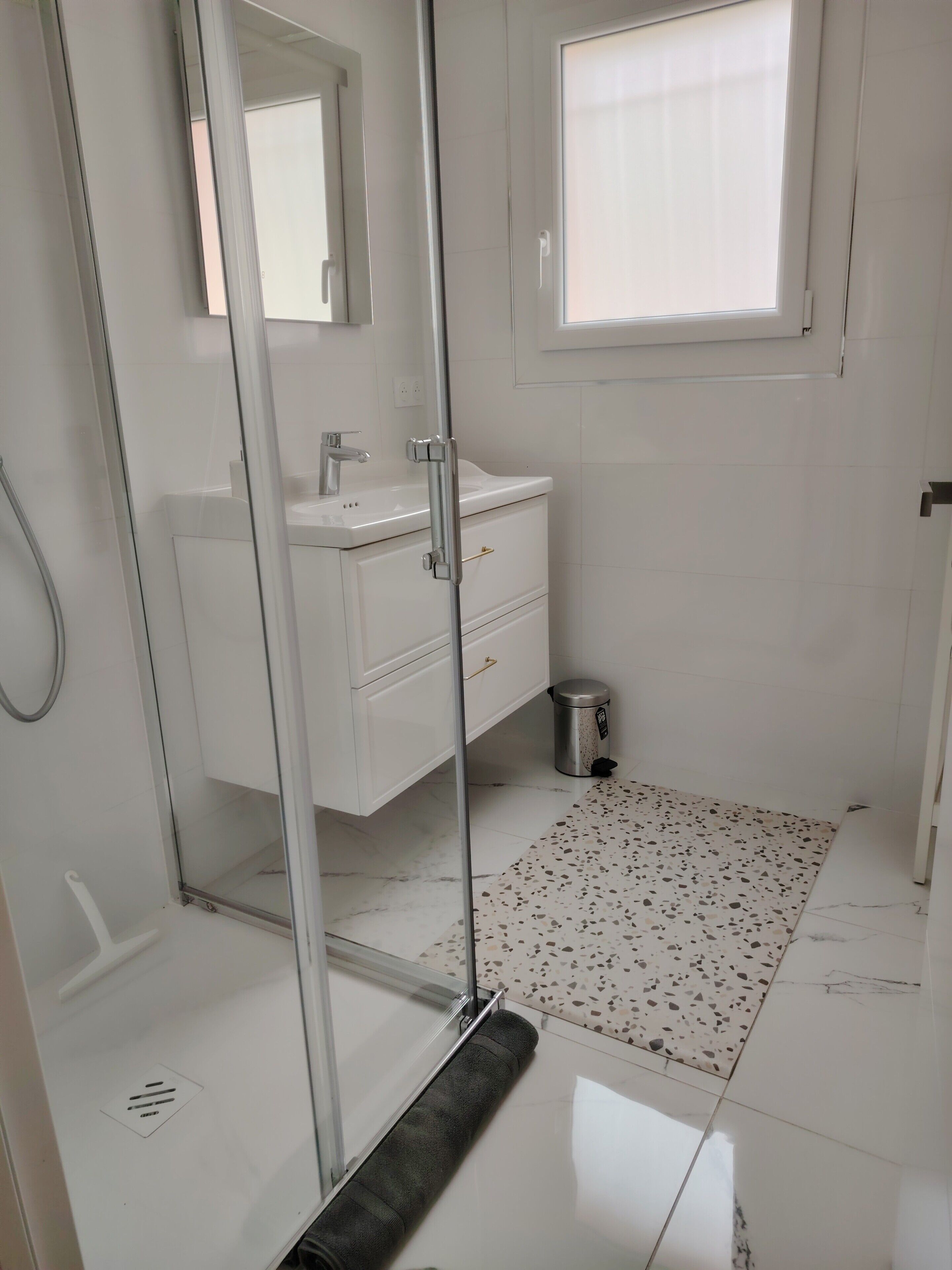 A bathroom with a glass shower enclosure, a white sink, a mirror, a window, and a tiled floor.