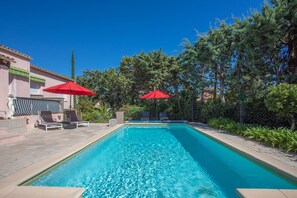 Una piscina rettangolare con acqua cristallina, circondata da lettini e ombrelloni rossi, con alberi e un edificio sullo sfondo.