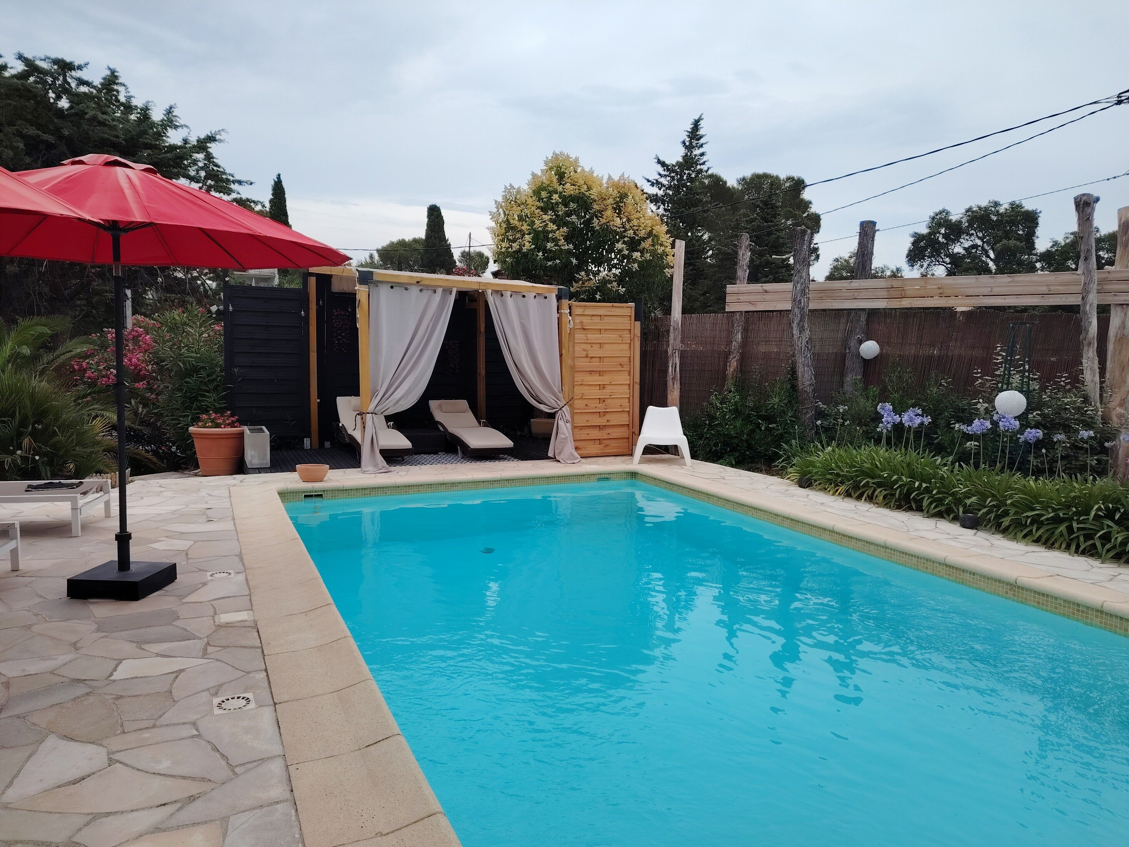 A swimming pool with a wooden deck, a red umbrella, and a covered seating area with white cushions.