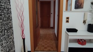 A hallway with a patterned floor, a wooden door, a wall with a tapestry, and a white shelf with items.