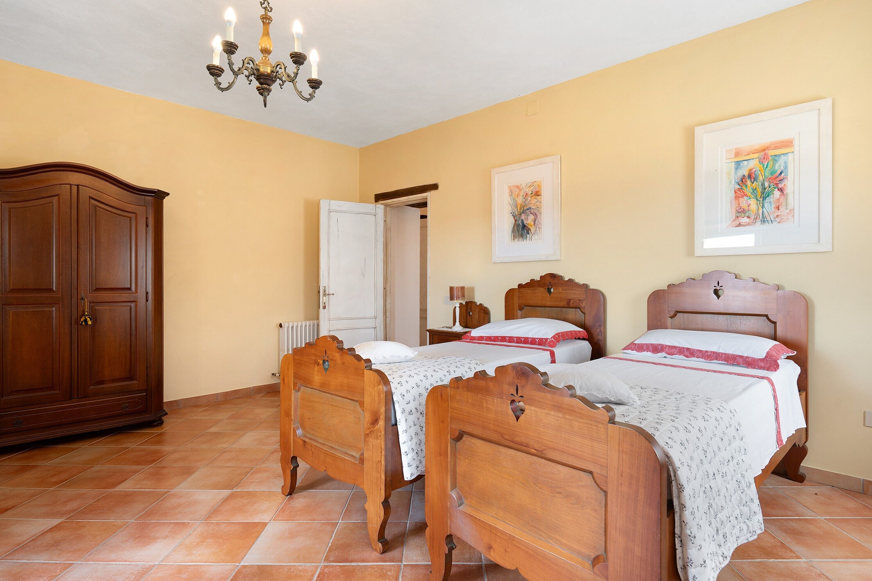 Un dormitorio con dos camas de madera, un armario, una araña y dos cuadros en las paredes.