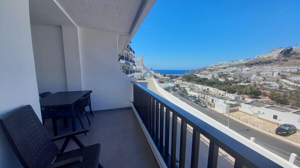 A balcony with a table and chairs, overlooking a coastal town with buildings and a clear blue sky.