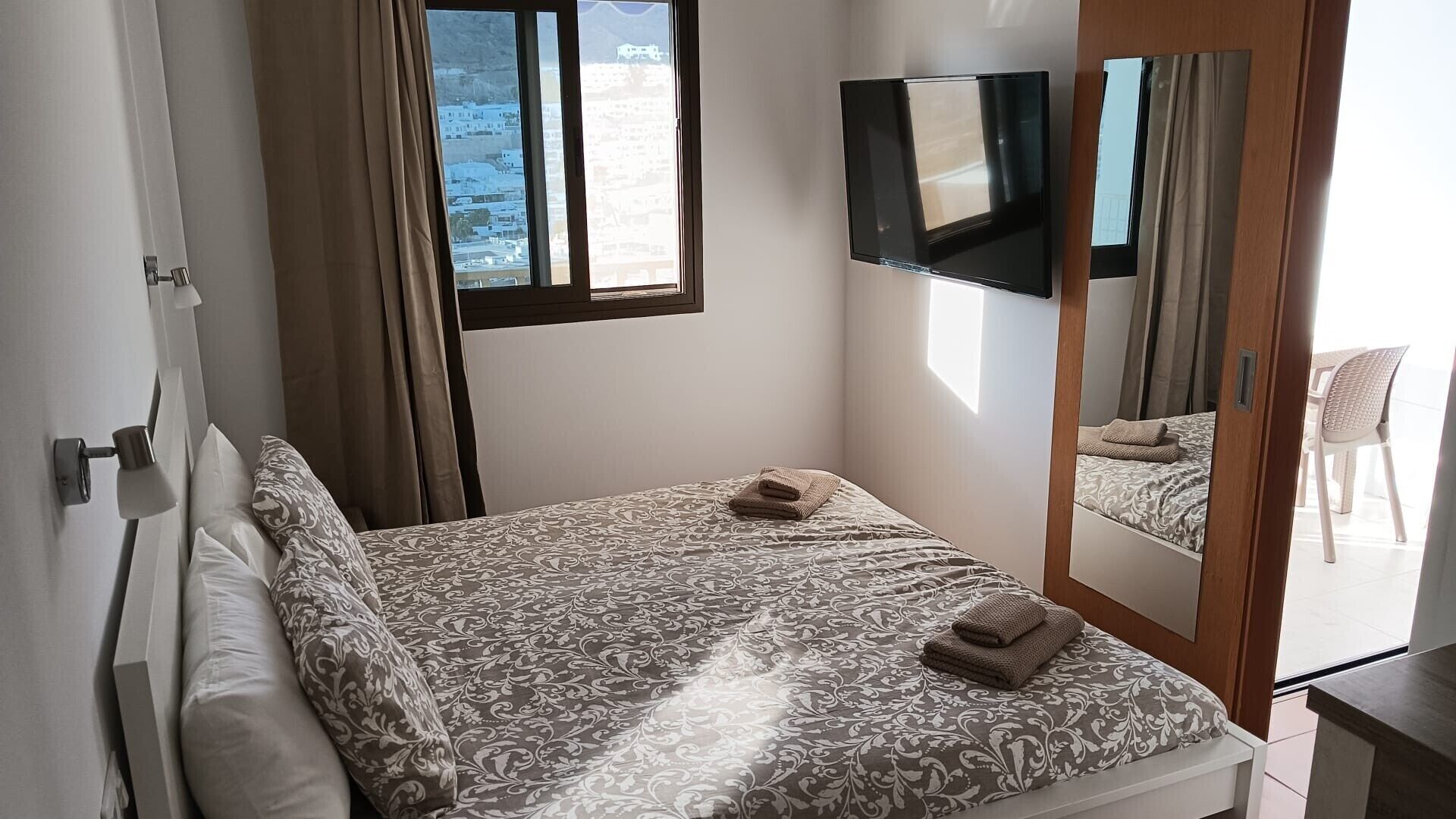 A bedroom with a bed, a TV, a mirror, and a window with a view of buildings.