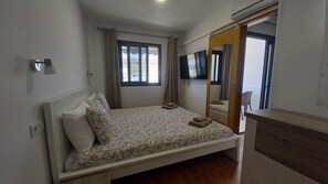 A single bed with patterned bedding, a bedside table, a mirror, a TV mounted on the wall, and a window with curtains.