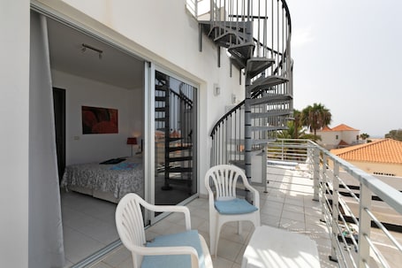 A balcony with a bed, a chair, a table, and a spiral staircase.