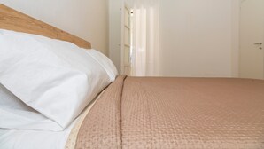 Une chambre à coucher avec une tête de lit en bois, un lit recouvert d’une literie blanche et beige, une porte blanche et une fenêtre ornée de rideaux légers.