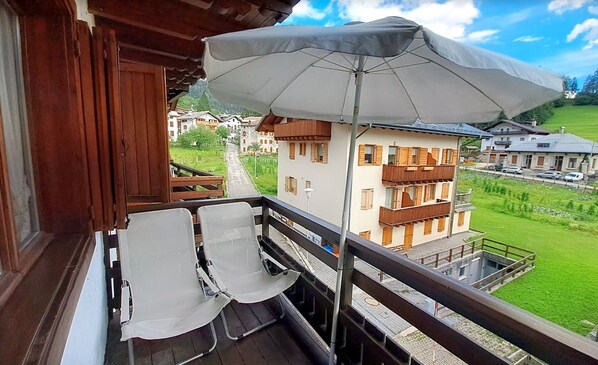 Un balcon avec deux chaises blanches, un grand parasol et une vue sur une ville avec ses bâtiments et sa verdure.