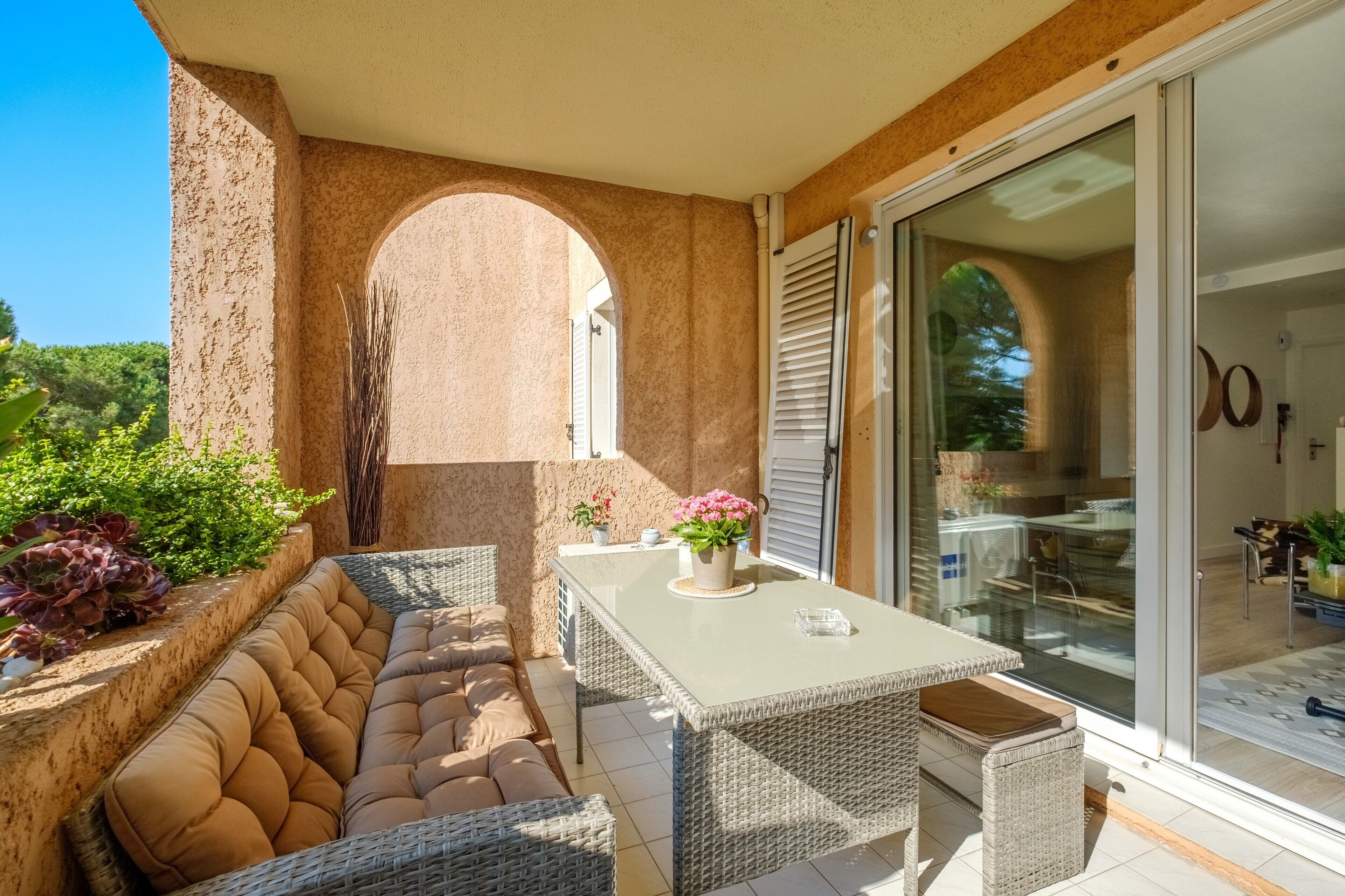 Une terrasse ensoleillée avec un coin salon confortable, une table agrémentée d’un vase de fleurs et une porte-fenêtre coulissante donnant sur l’intérieur.
