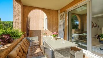 Une terrasse ensoleillée avec un coin salon confortable, une table agrémentée d’un vase de fleurs et une porte-fenêtre coulissante donnant sur l’intérieur.