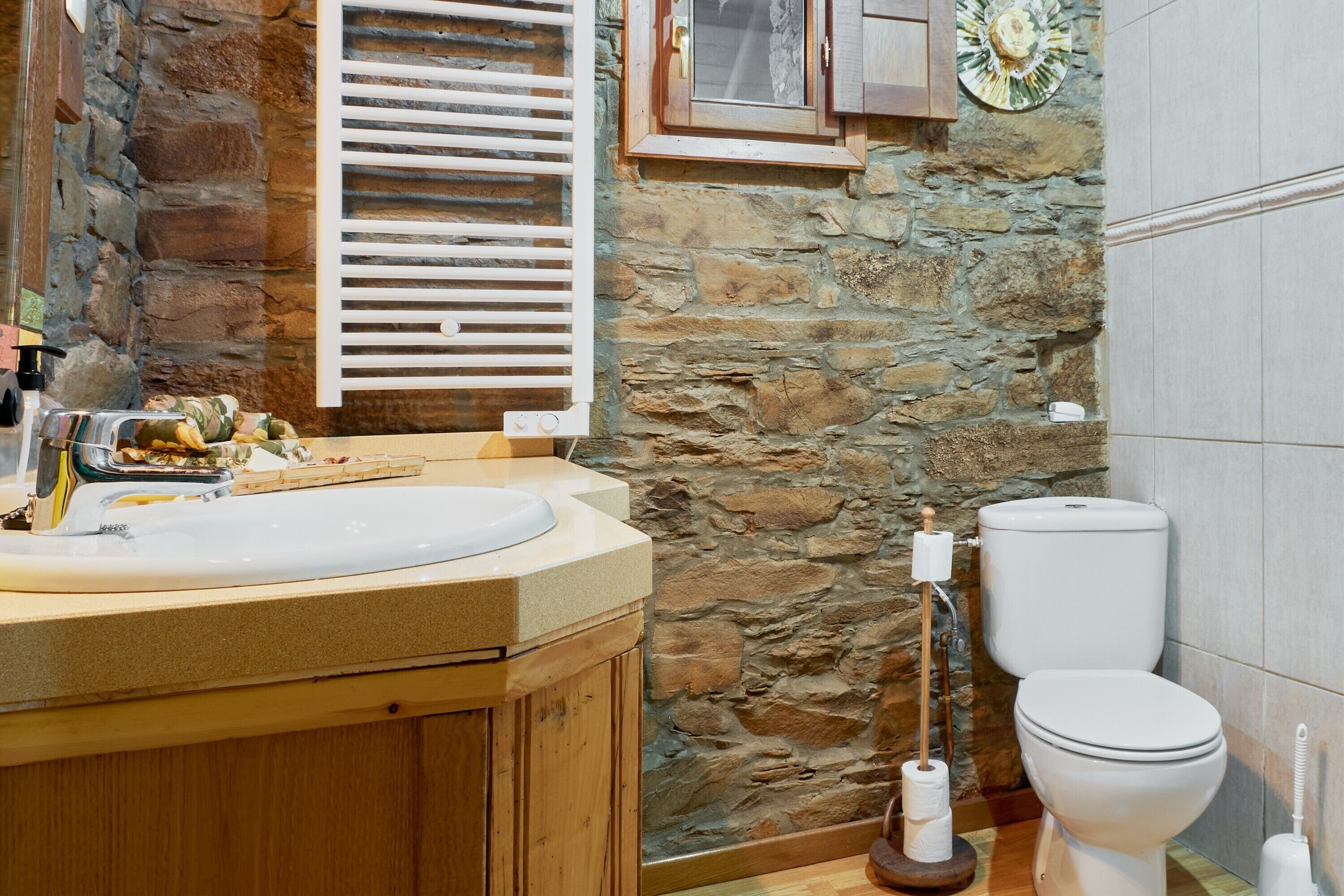 Un baño con pared de piedra, armarios de madera, lavamanos, inodoro y una ventana con contraventanas.