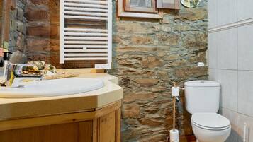 Un baño con pared de piedra, armarios de madera, lavamanos, inodoro y una ventana con contraventanas.