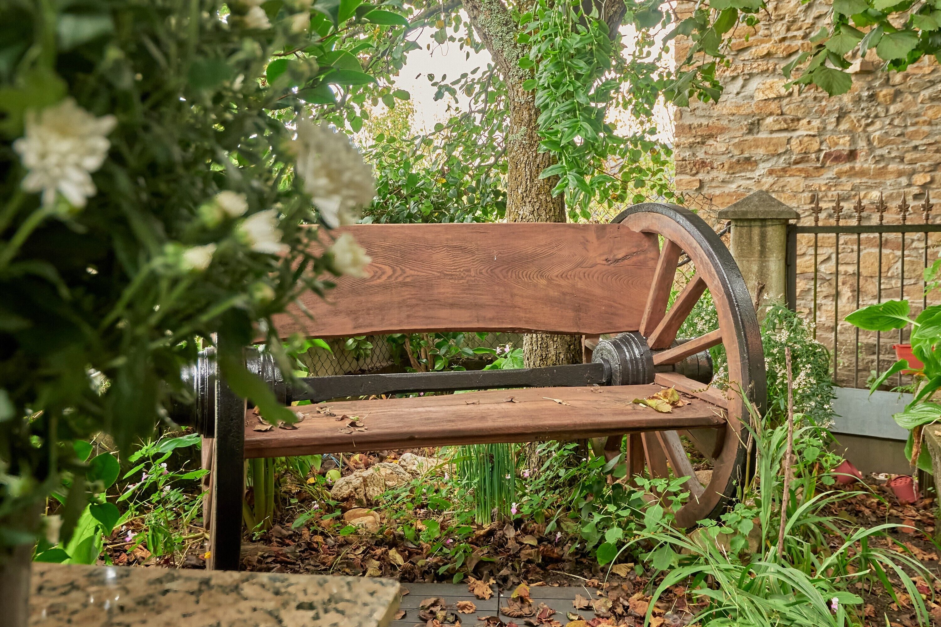 A wooden bench with a large wheel, surrounded by greenery and a stone wall.