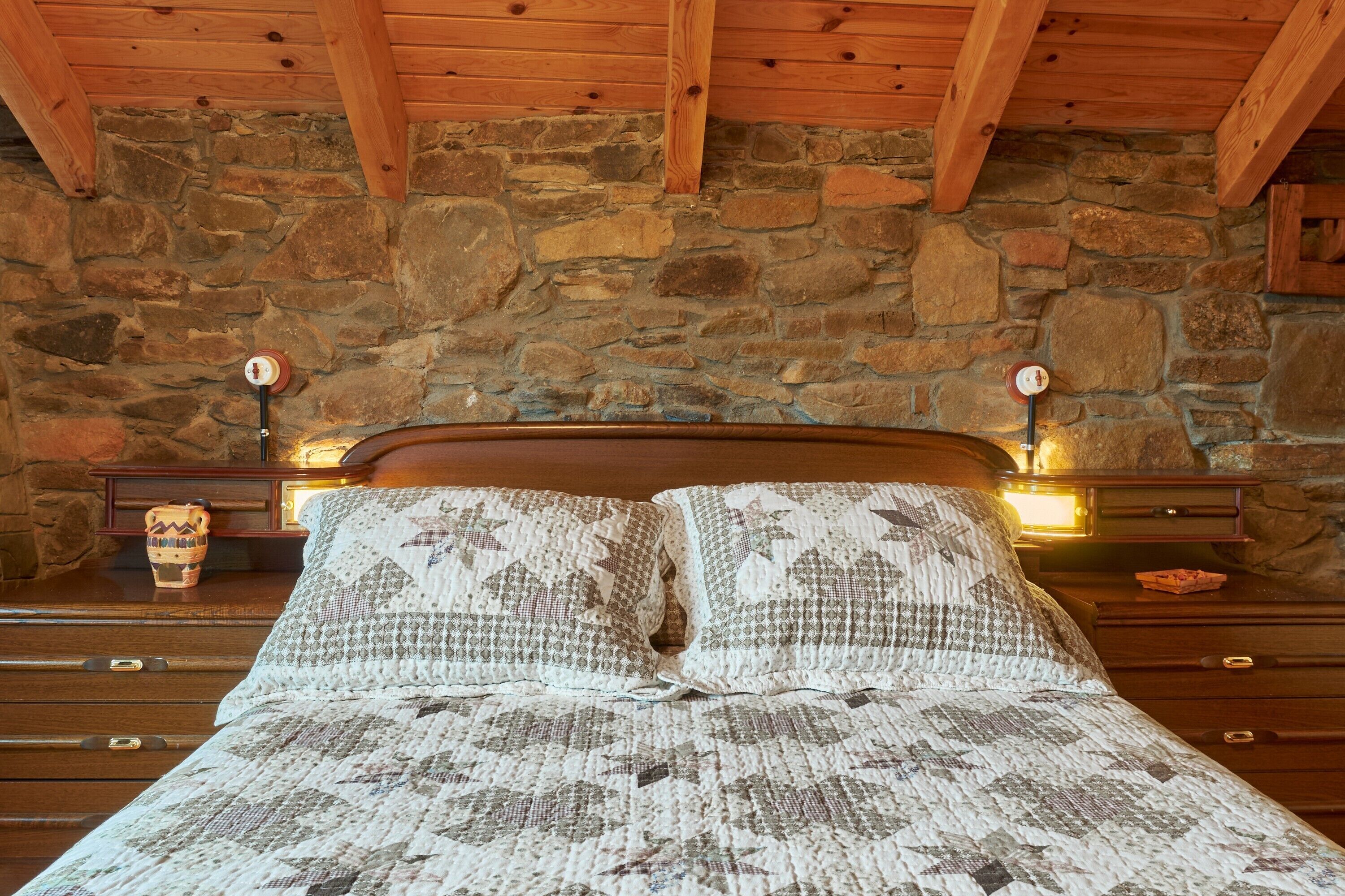 Ein Bett mit gemusterten Betttextilien, einem Holzkopfende, einer Steinwand und Nachttischlampen.