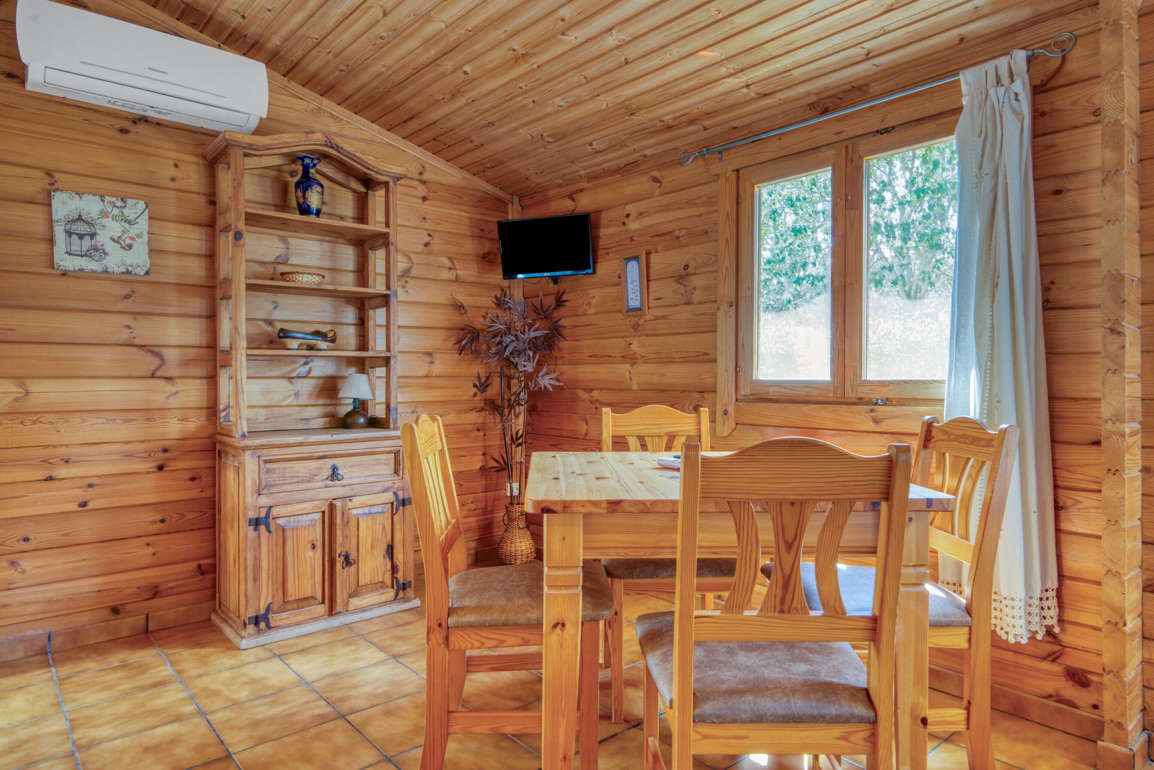Interior de una cabaña de madera con una mesa de comedor, sillas, un televisor montado en la pared y una ventana con cortinas.