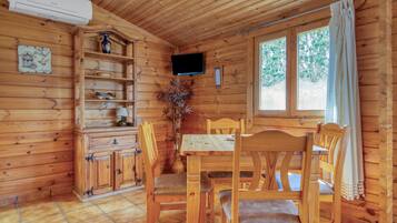 Interior de una cabaña de madera con una mesa de comedor, sillas, un televisor montado en la pared y una ventana con cortinas.