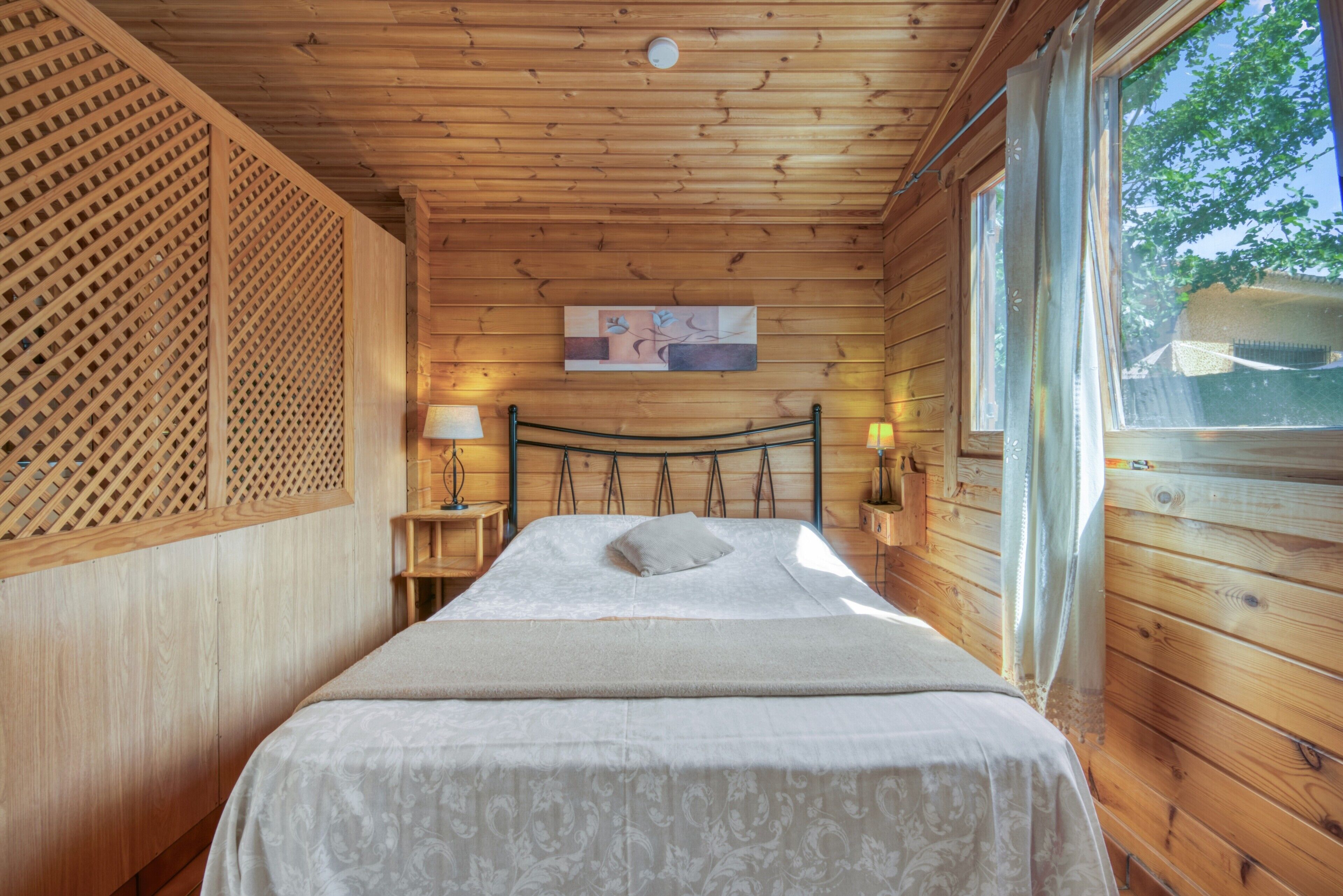 Un dormitorio de cabaña de madera con una cama, una mesita de noche, una lámpara y un ventanal.