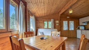 A wooden cabin interior with a dining table, chairs, a kitchen area, and large windows.