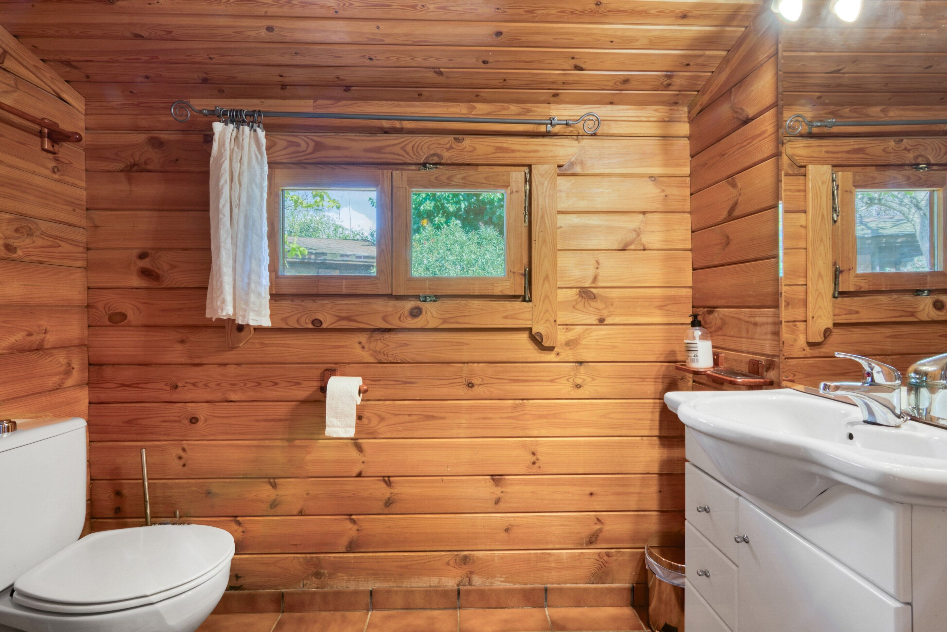 Un baño rústico en una cabaña de madera con inodoro, lavamanos y dos ventanas.