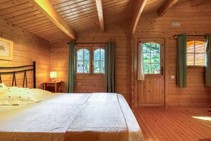 A wooden cabin room with a bed, a small table, a lamp, and two windows with curtains.