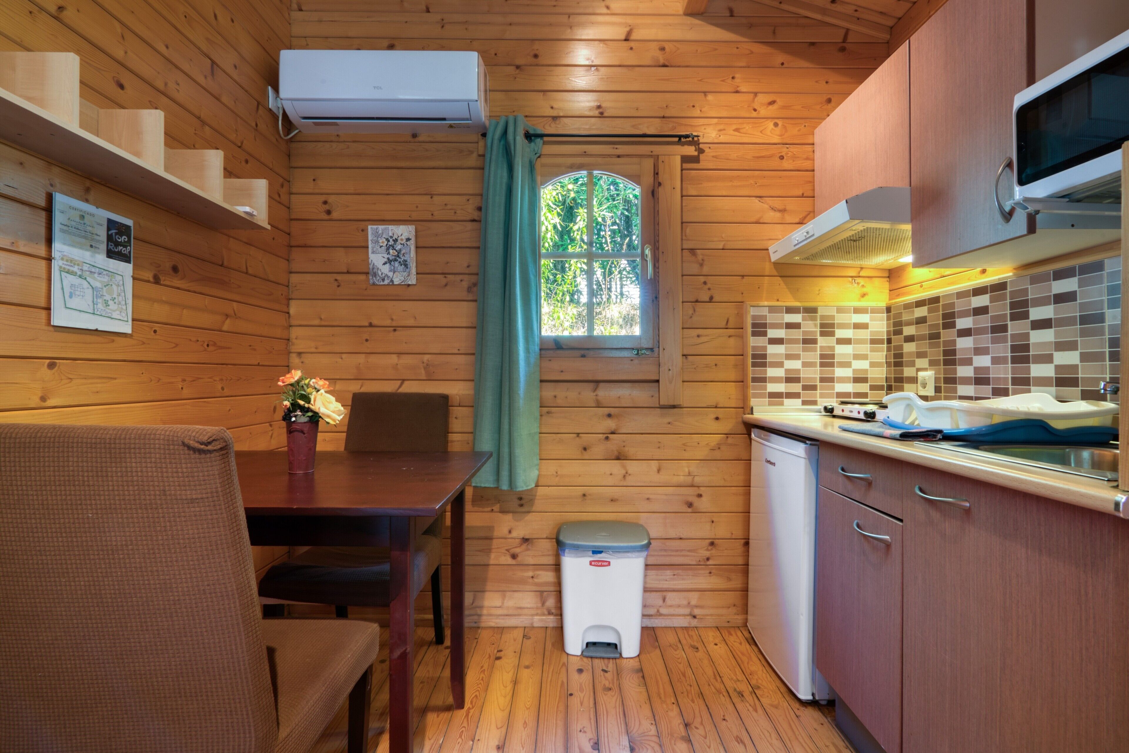 A wooden cabin interior with a dining area, kitchen, and a window with curtains.