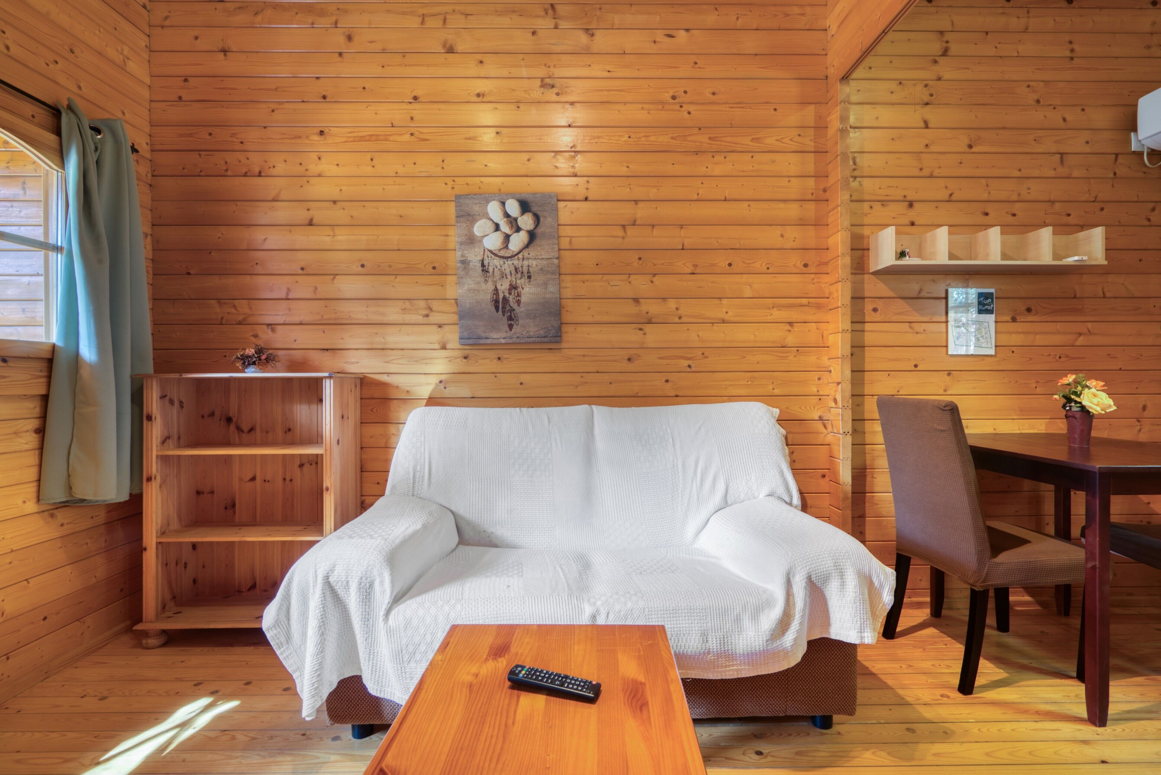 Interior de una cabaña de madera con un sofá blanco, una mesa de centro de madera, una mesa de comedor con sillas y una estantería de madera con un cuadro.