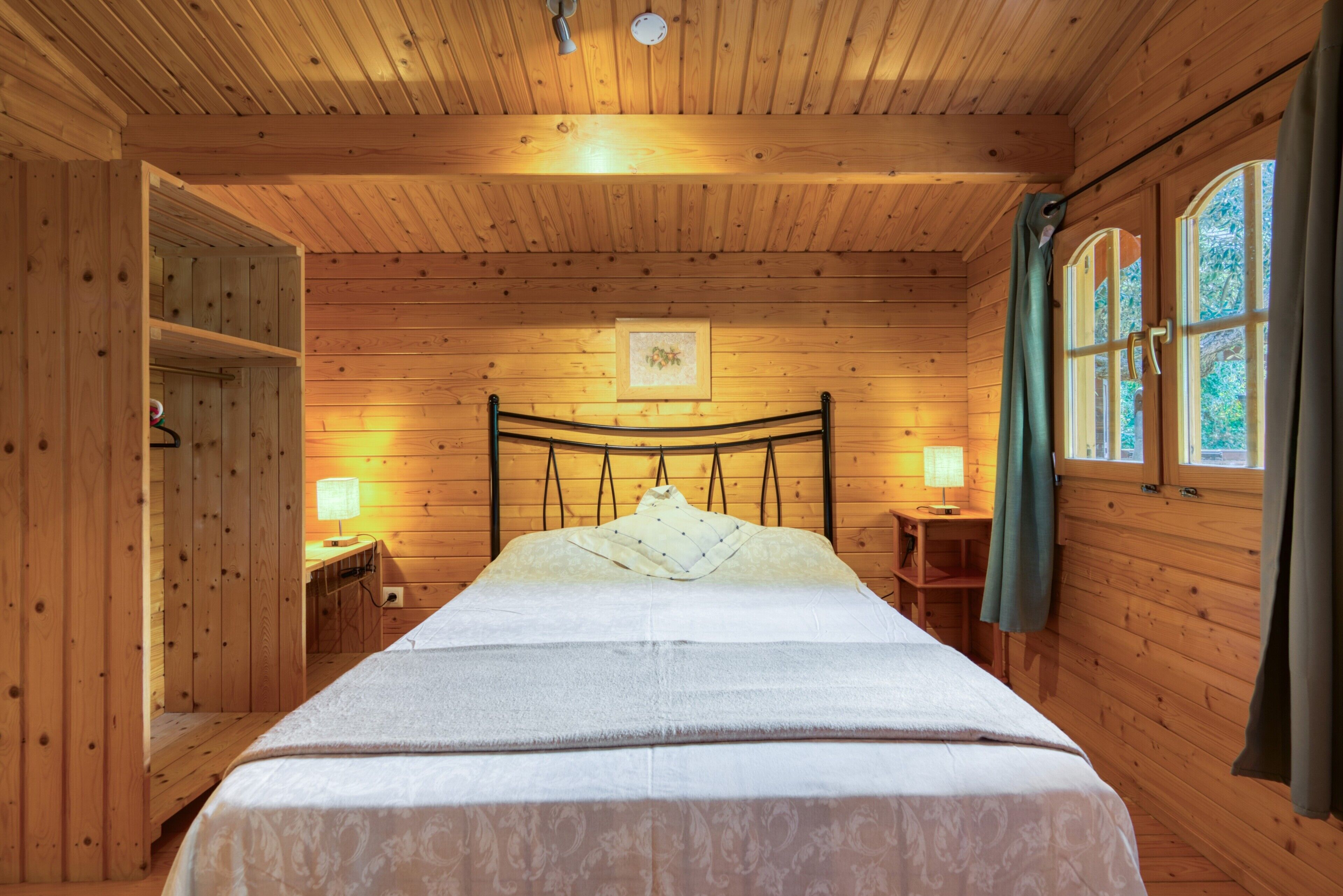 A wooden cabin bedroom with a bed, bedside lamps, a wooden wardrobe, and a window with curtains.