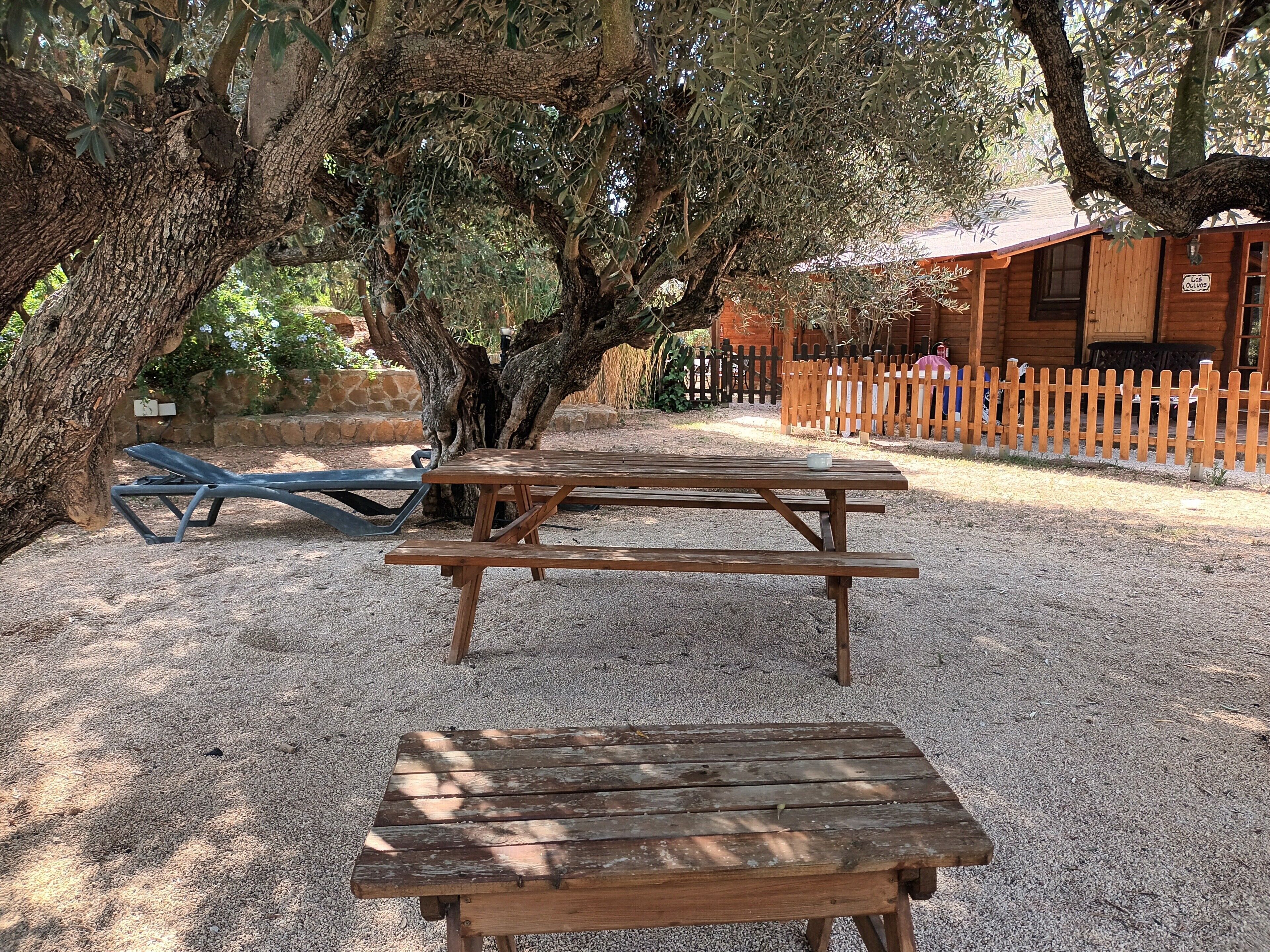 A wooden picnic table and lounge chair in a gravel area with trees and a wooden fence.