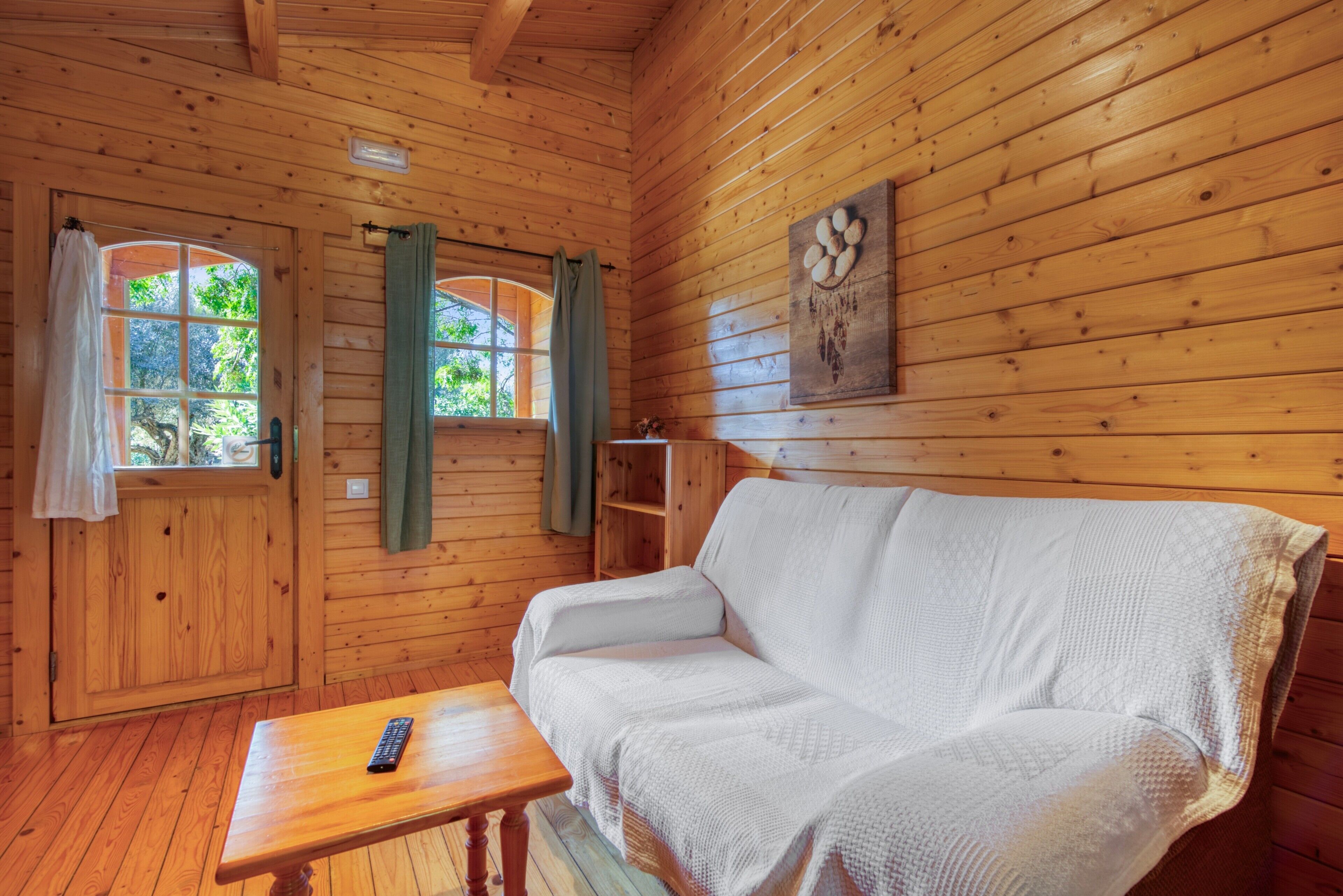 Interior de una cabaña de madera con sofá, mesa y ventana con cortinas.