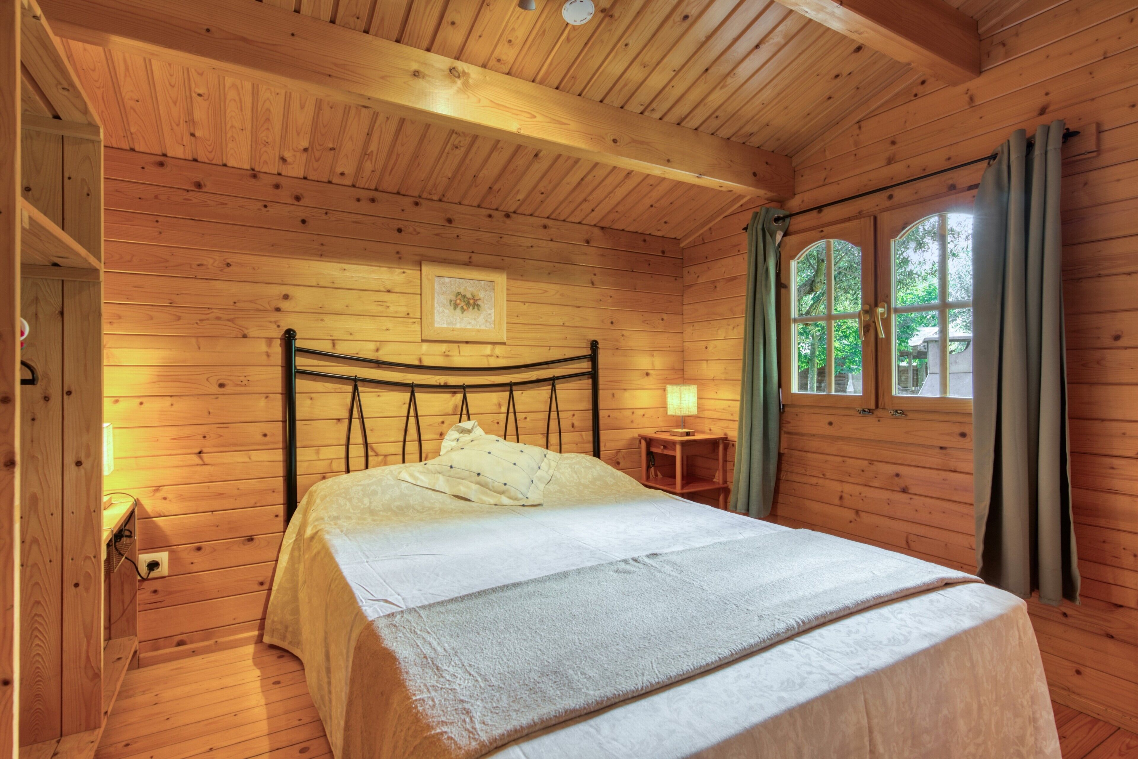 A wooden cabin bedroom with a bed, a nightstand, a window with curtains, and a framed picture on the wall.