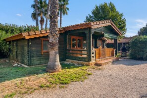 A rustic wooden cabin with a porch, surrounded by greenery and a gravel path.