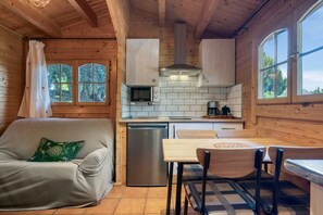 Et interiør af en træhytte med et tekøkken, en sofa med en grøn pude, et spisebord med stole og udsigt til træer gennem et vindue.