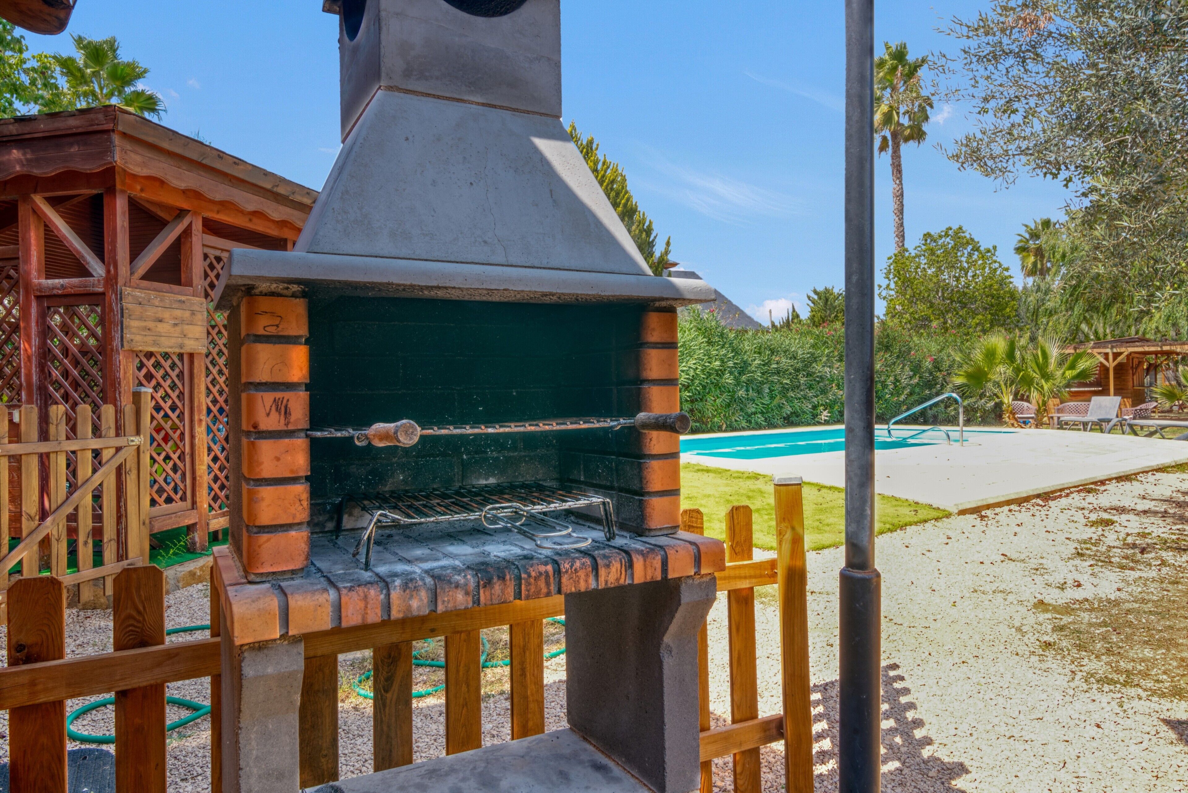 A brick-built outdoor grill with a chimney, surrounded by a wooden fence and a pool in the background.