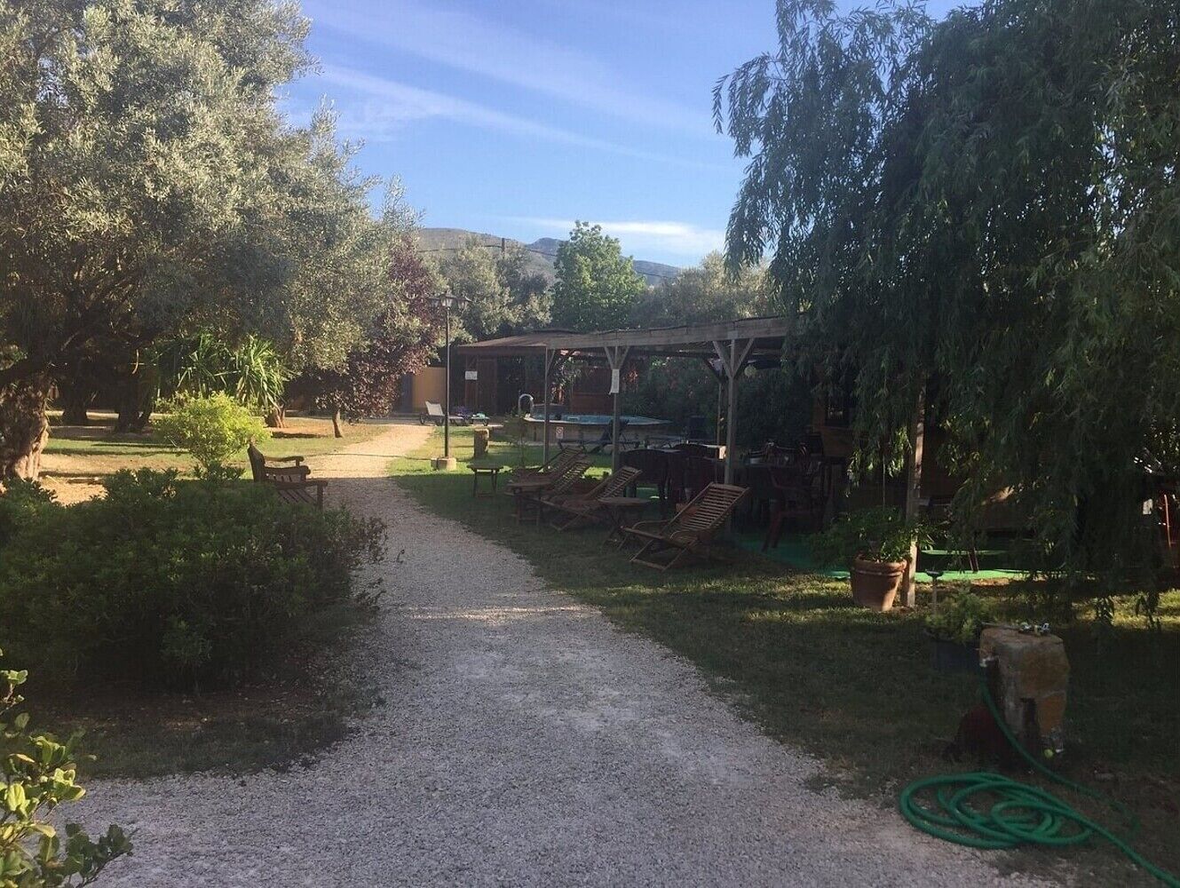A gravel path leading to a covered outdoor seating area with chairs and tables, surrounded by trees and a building in the background.