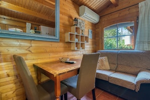A wooden cabin interior with a dining table, chairs, a sofa, and a window with a view of greenery.