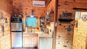 Cocina compacta con paredes de madera, un refrigerador de acero inoxidable, un lavamanos y una ventana con cortina de cuadros.