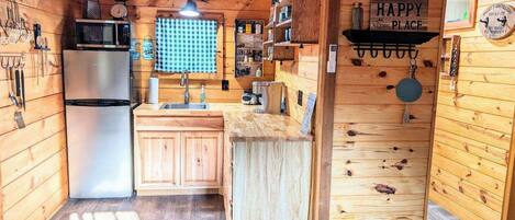 Cocina compacta con paredes de madera, un refrigerador de acero inoxidable, un lavamanos y una ventana con cortina de cuadros.