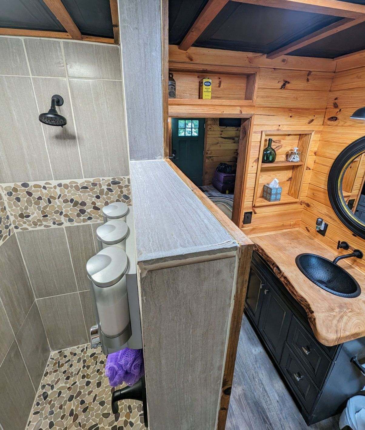 Een badkamer met een douche, wastafel en toilet. Er is een houten plank met toiletartikelen en een spiegel.