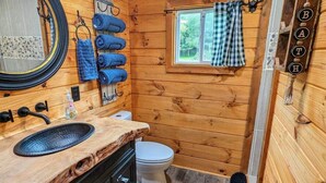 Un baño de cabaña de madera con un lavabo de madera, un inodoro y una ventana con cortina a cuadros.