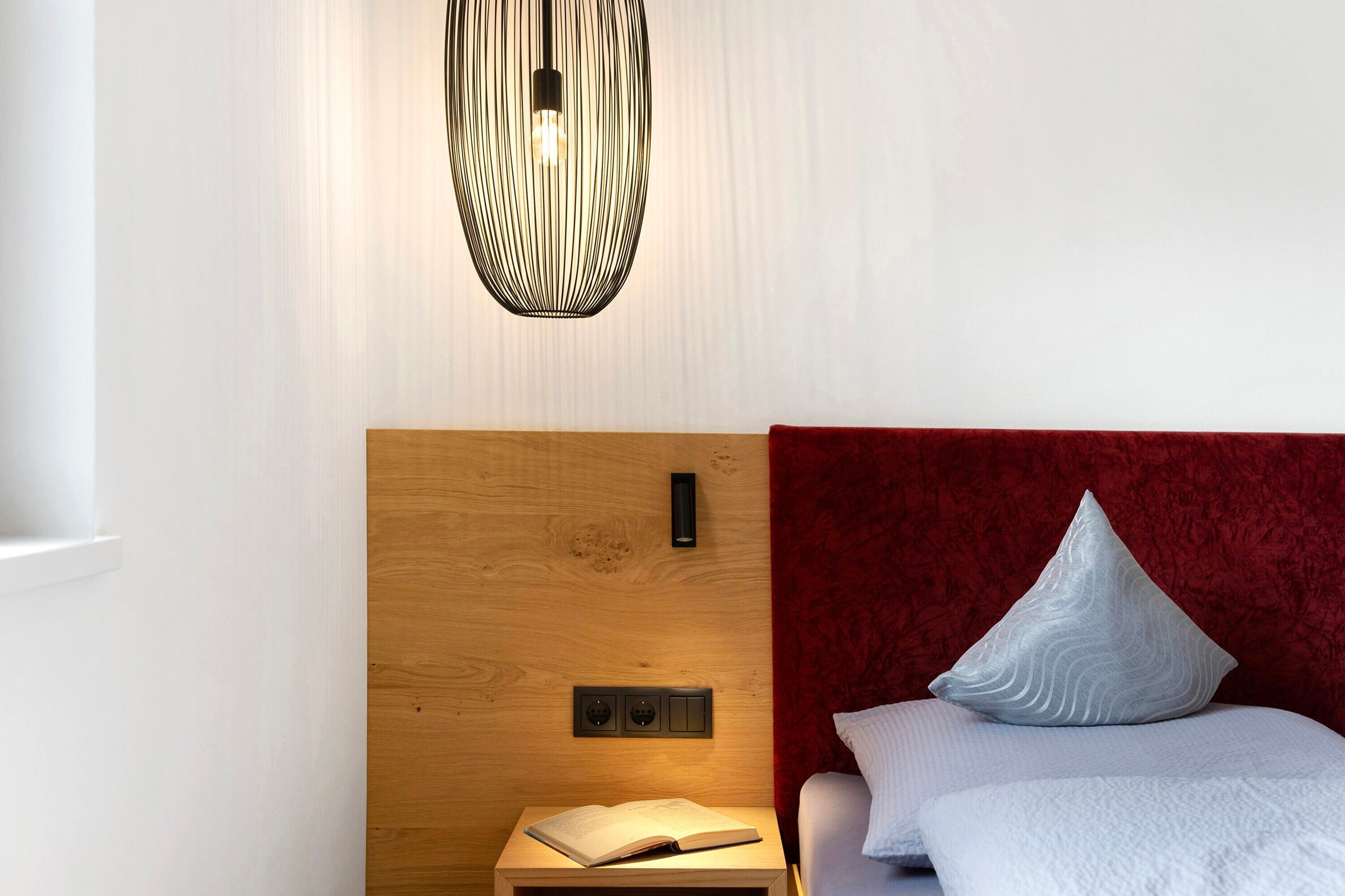 A neatly made bed with a red headboard, a bedside table with an open book, and a modern pendant light fixture.