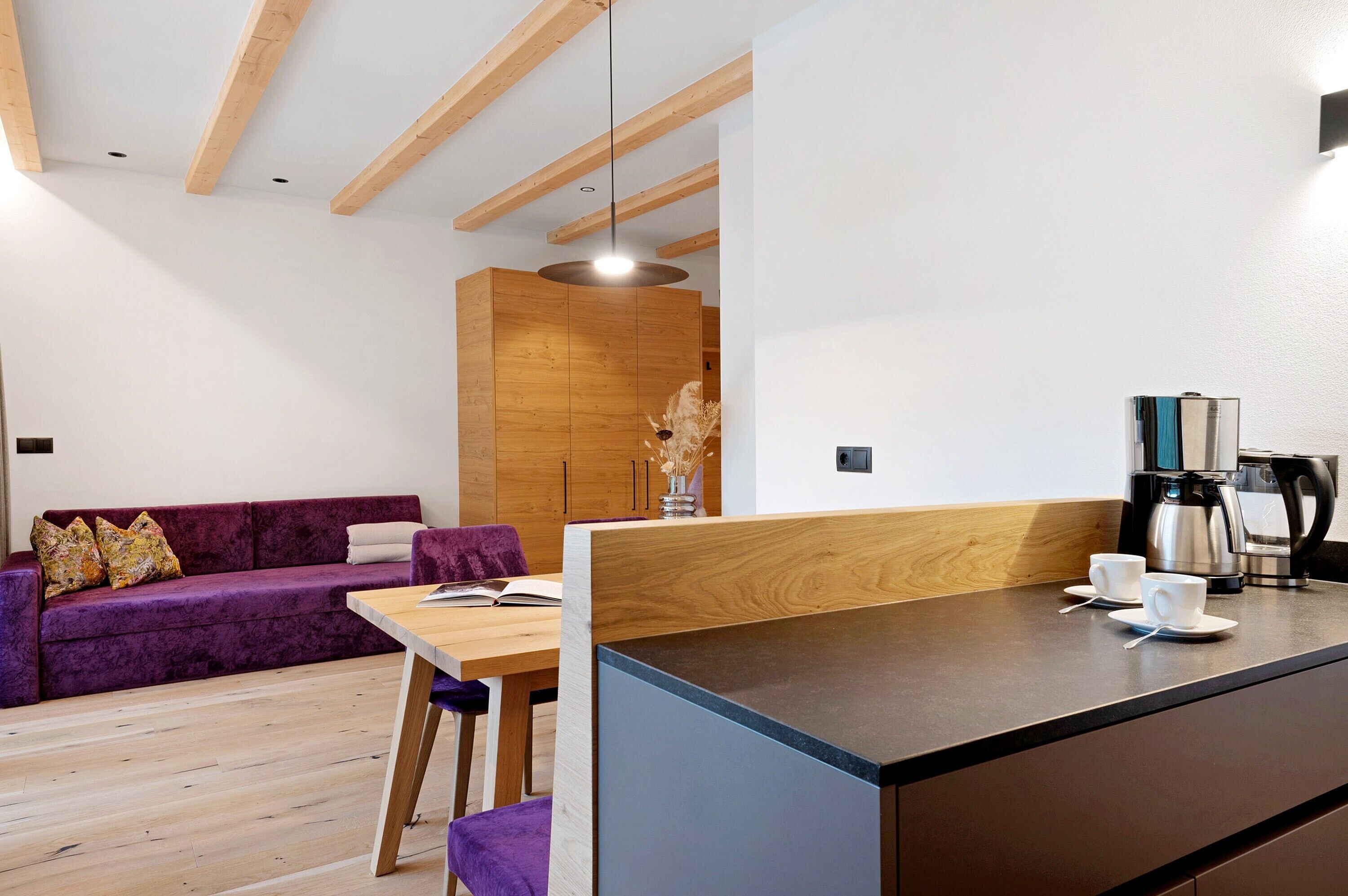 Ein modernes Interieur mit Holzboden, einem violetten Sofa, einem Holzesstisch und einer Kaffeemaschine.