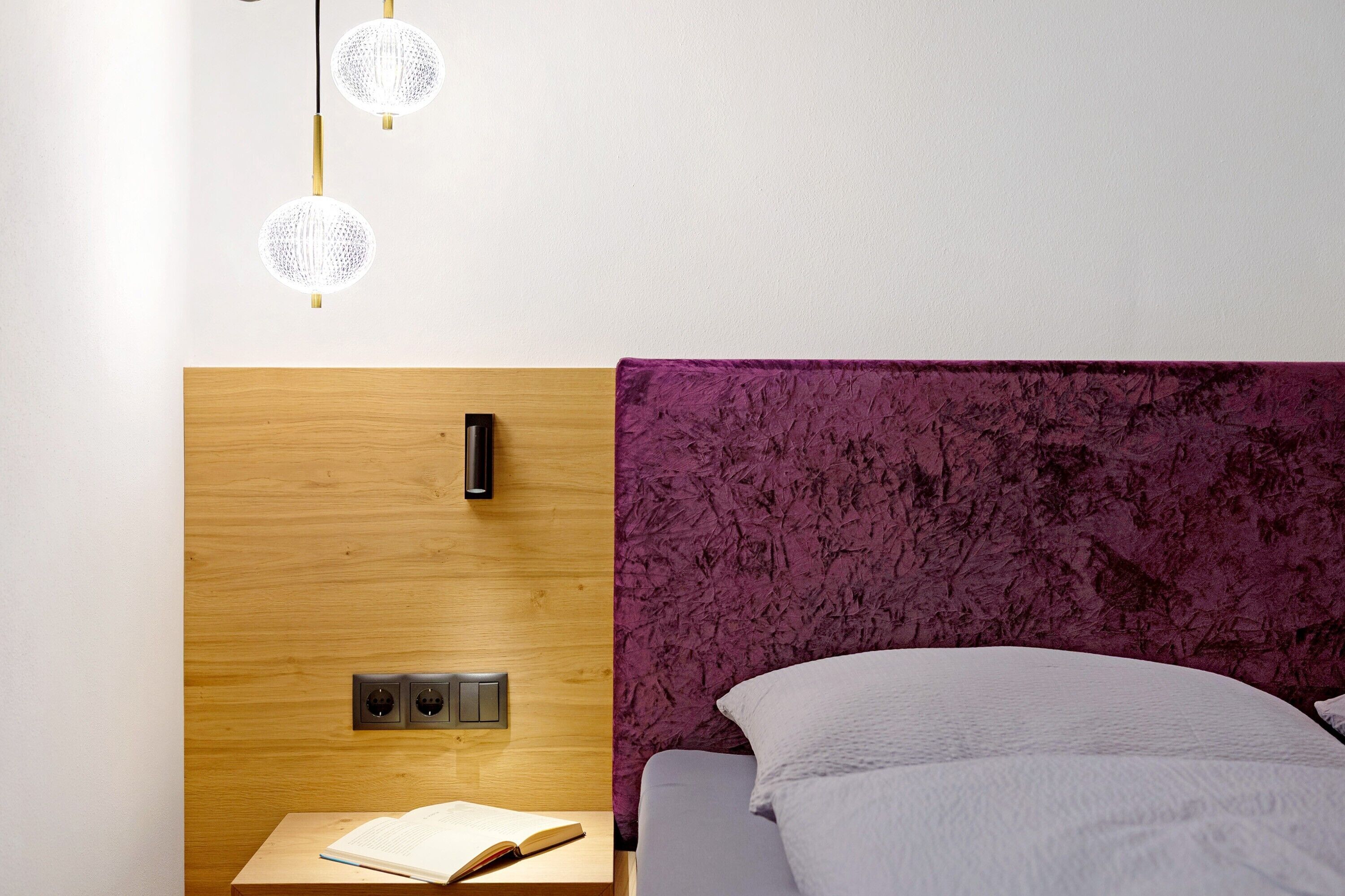 Una habitación con cabecera de madera, pared texturizada de color morado, cama con ropa de cama blanca, mesita de noche con un libro abierto y un aplique de pared.
