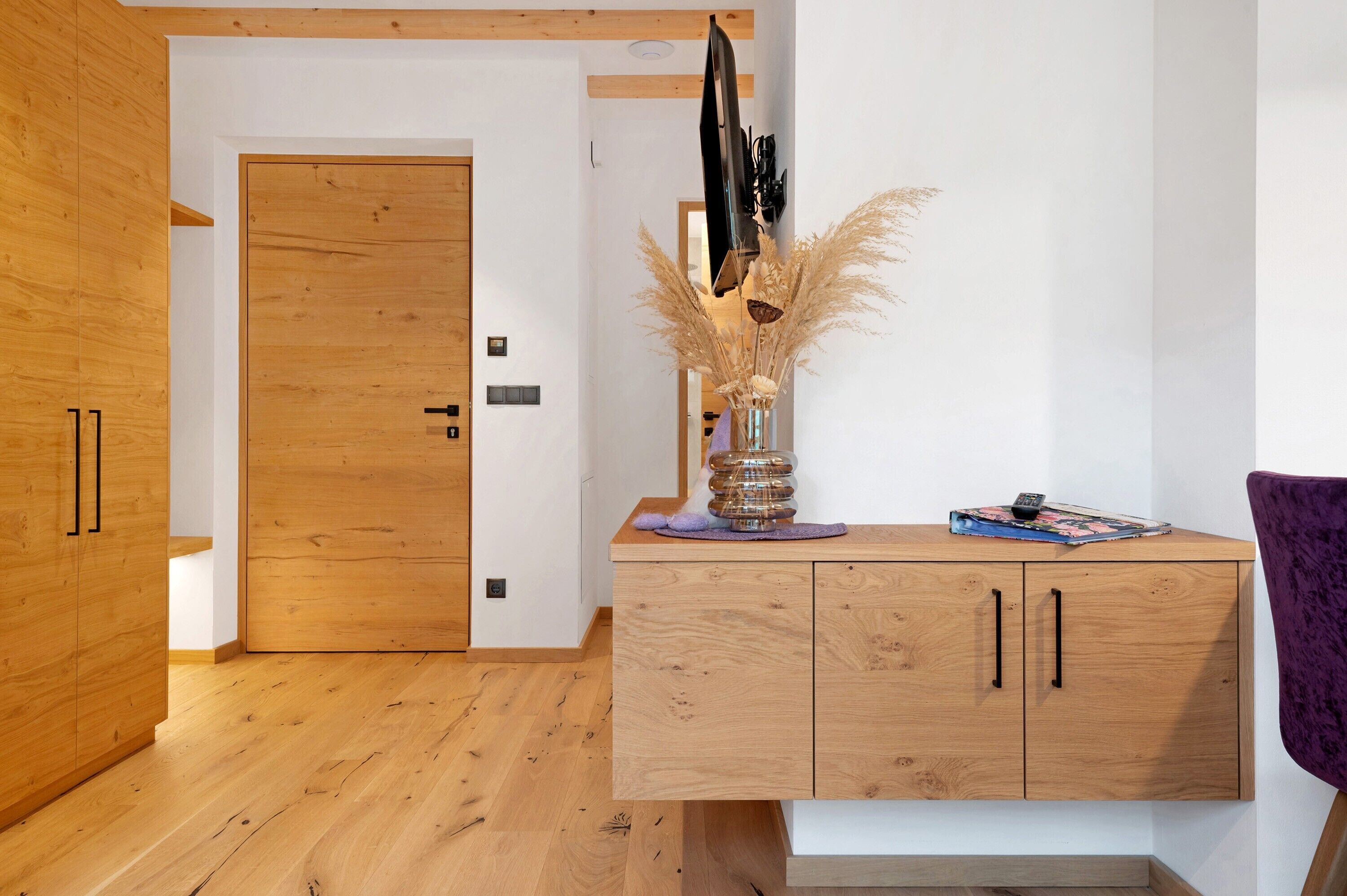 A wooden door, a wooden cabinet with two drawers, a vase with dried plants, and a purple chair.