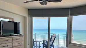Une chambre à coucher avec un lit, un téléviseur posé sur une commode, un ventilateur de plafond et un balcon offrant une vue sur l’océan.
