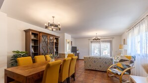 Un'area pranzo con tavolo in legno e sedie gialle, un divano con cuscini decorati e un lampadario.