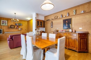 Une pièce aux murs lambrissés, comprenant une table à manger, des chaises, un canapé et un coin cuisine avec des placards et une horloge.