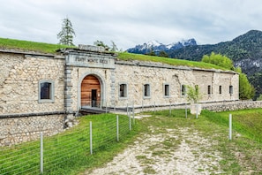 Ein Steinhaus mit einer Holztür, beschriftet mit 'FORT. MONTE RICCO', umgeben von einer grünen Fläche und Bergen im Hintergrund.